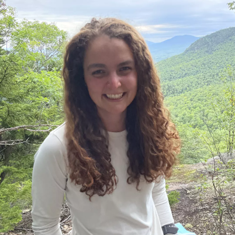The image shows a young woman with curly brown hair smiling at the camera. She is standing outdoors, with a scenic mountain view in the background. She is wearing a light-colored long-sleeved shirt. The overall impression is one of happiness and connection with nature.